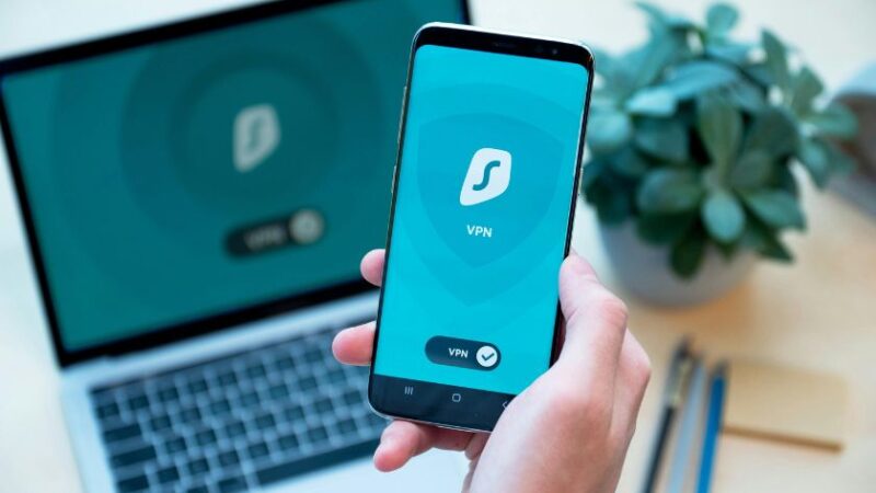 Une main tenant un smartphone affichant une application VPN avec un ordinateur portable en arrière plan montrant le même écran