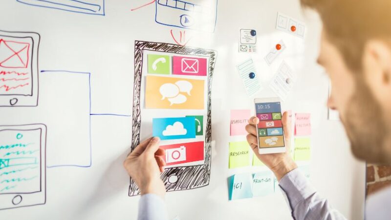 Designer travaillant sur un tableau blanc avec maquettes dapplication mobile et schémas UX
