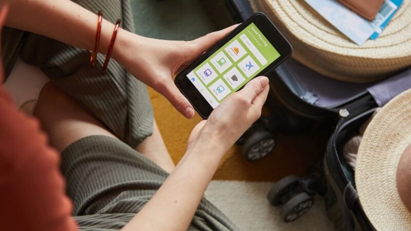 Femme utilisant une application mobile de voyage sur smartphone assise à côté dune valise ouverte avec passeport et chapeau de paille